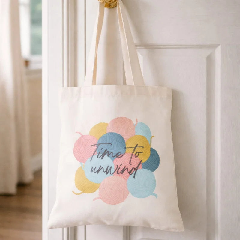 An off-white canvas tote bag hanging on a white door, featuring a graphic of pastel-colored yarn balls with the phrase 'Time to unwind' written across them.