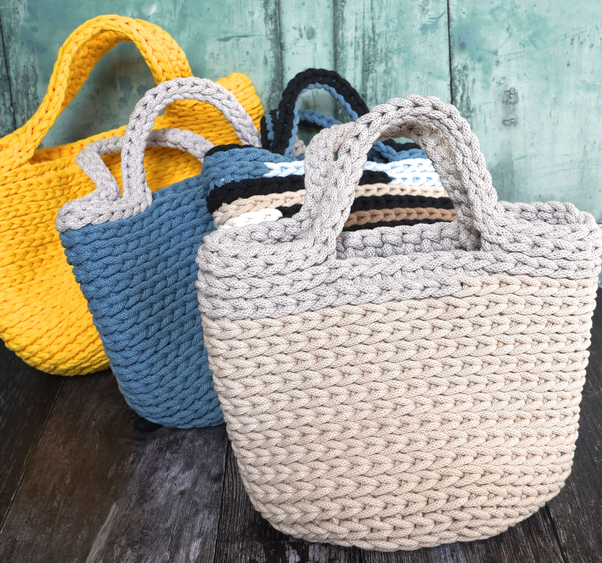 Three crocheted bags in yellow, blue, and beige on a wooden surface with a teal wooden background.