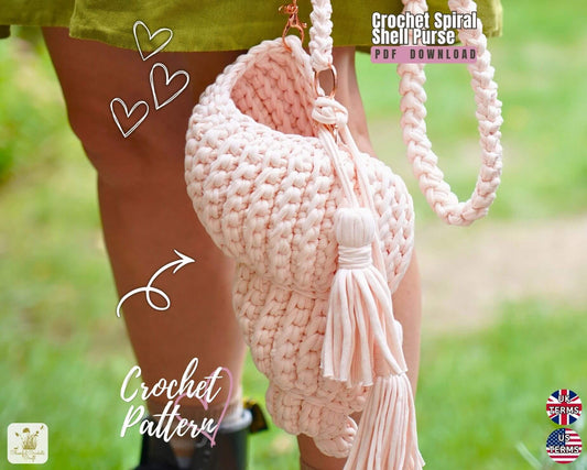 A crocheted shell-shaped shoulder bag in pale blush pink, shown being worn outdoors with a person's legs visible. The bag has a chunky spiral crochet texture, a braided shoulder strap, and a large tassel.