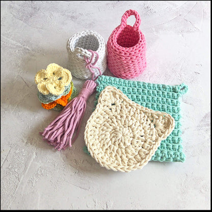 Collection of crocheted items including a pink basket, a beige dishcloth, a green scrubber, and a tassel on a light gray surface.