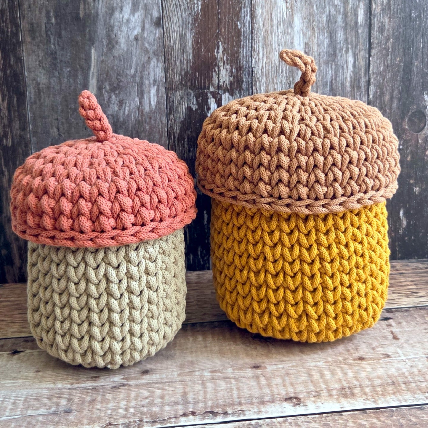 Two crocheted acorn-shaped containers with textured yarn bodies and wooden-look lids, displayed on a rustic wooden surface against a weathered wood background.
