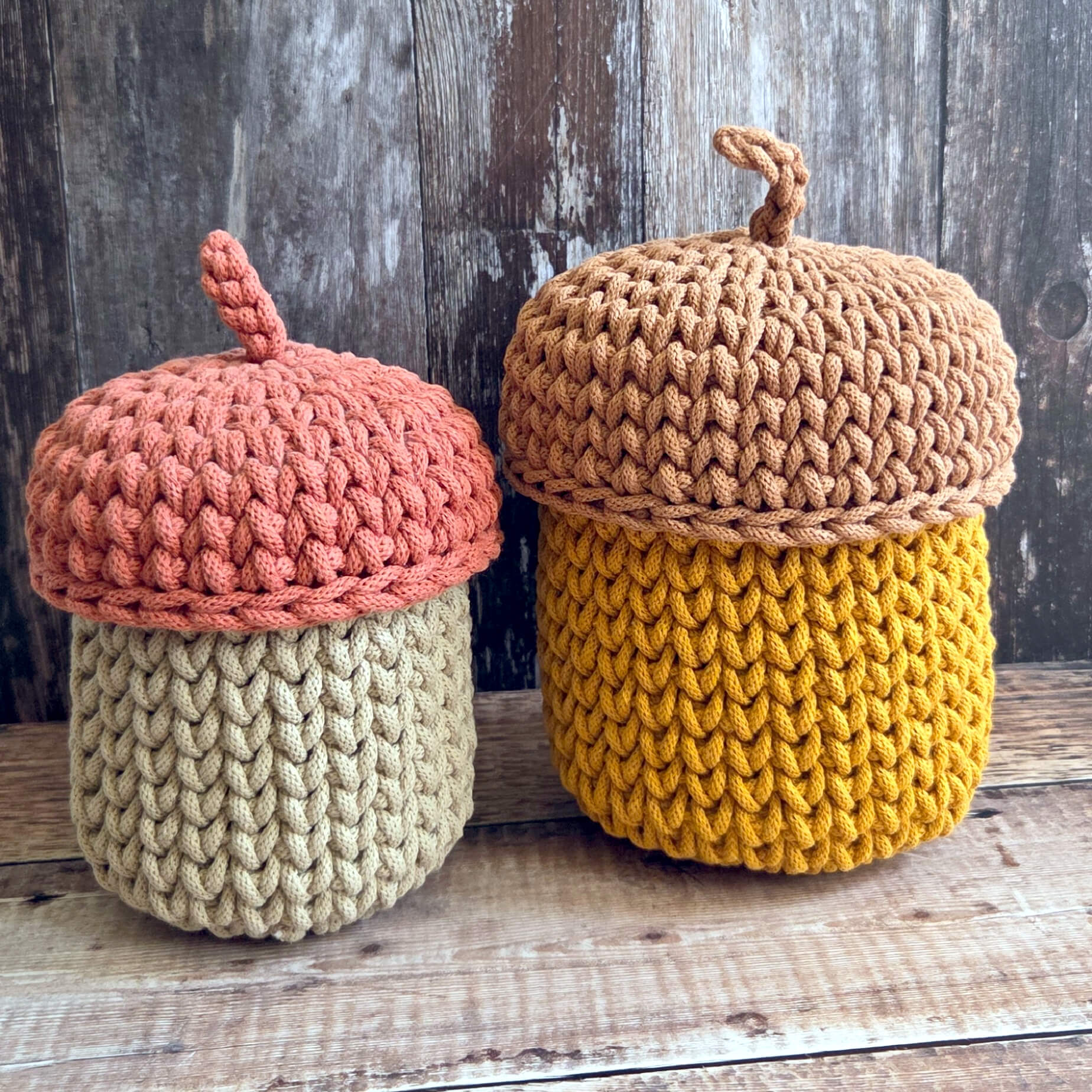 Two crocheted acorn-shaped containers with textured yarn bodies and wooden-look lids, displayed on a rustic wooden surface against a weathered wood background.