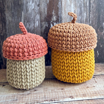 Two crocheted acorn-shaped containers with textured yarn bodies and wooden-look lids, displayed on a rustic wooden surface against a weathered wood background.
