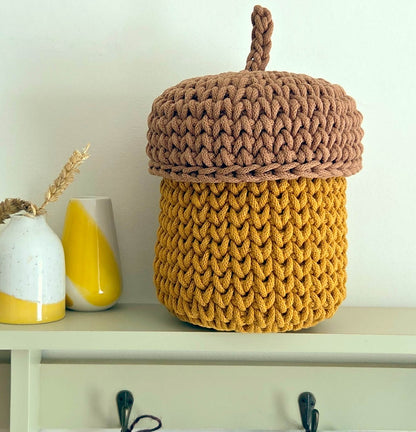 A chunky knit acorn-shaped storage basket with a brown top and handle and a mustard yellow body, displayed on a shelf next to two ceramic vases.