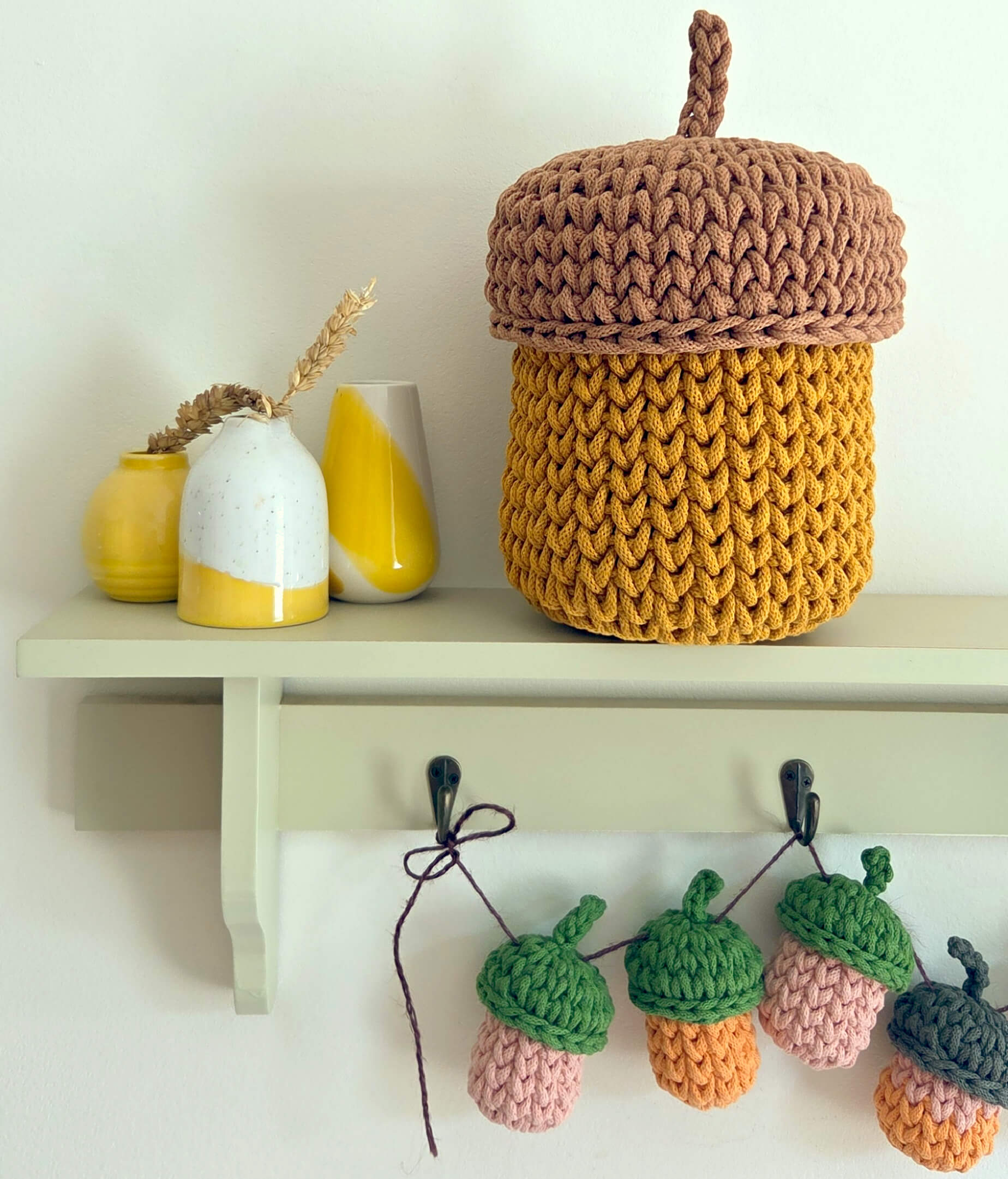 A large crocheted acorn-shaped storage basket in brown and mustard yellow sits on a light green shelf, with smaller decorative vases on the left and matching crocheted acorns hanging from black hooks below.