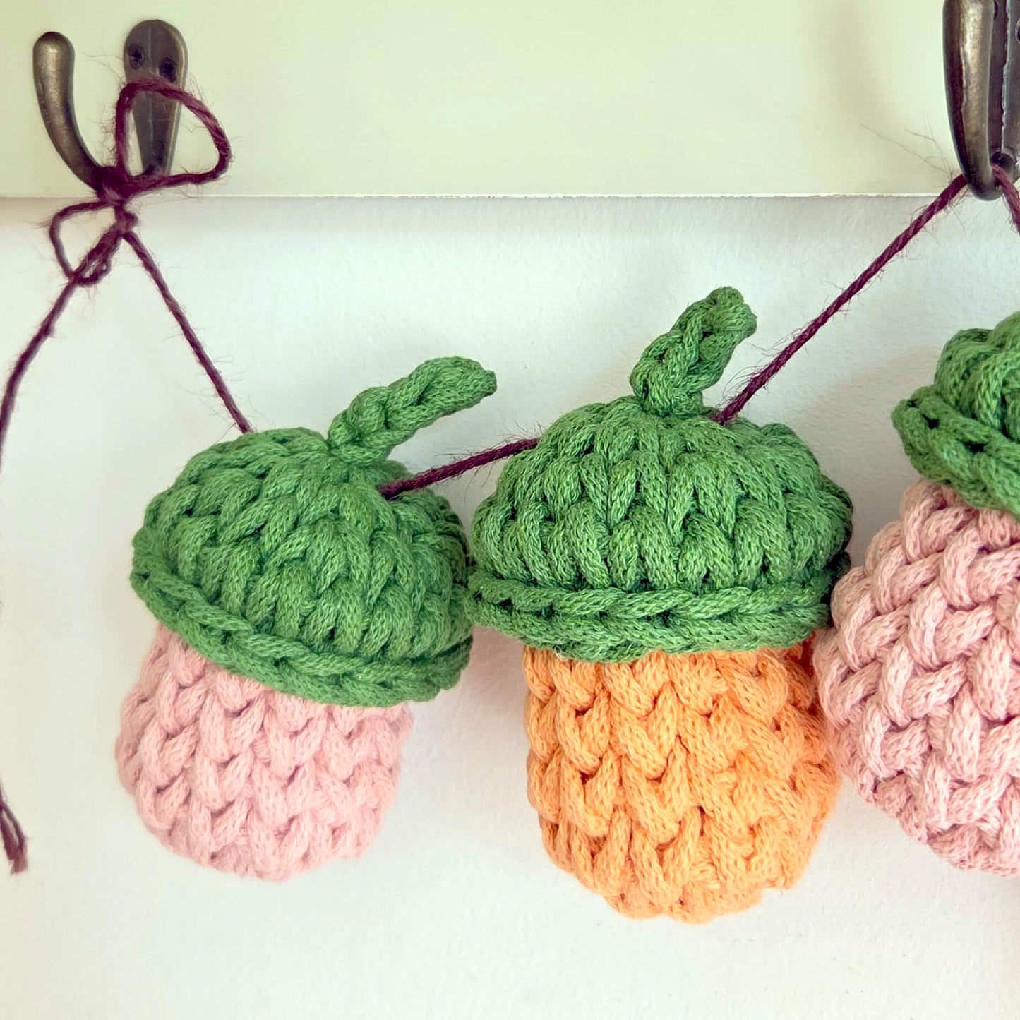 Three crocheted acorn ornaments hanging from dark hooks against a plain light background. The acorns have green textured caps, pink and orange textured bodies, and are strung on dark burgundy cords.