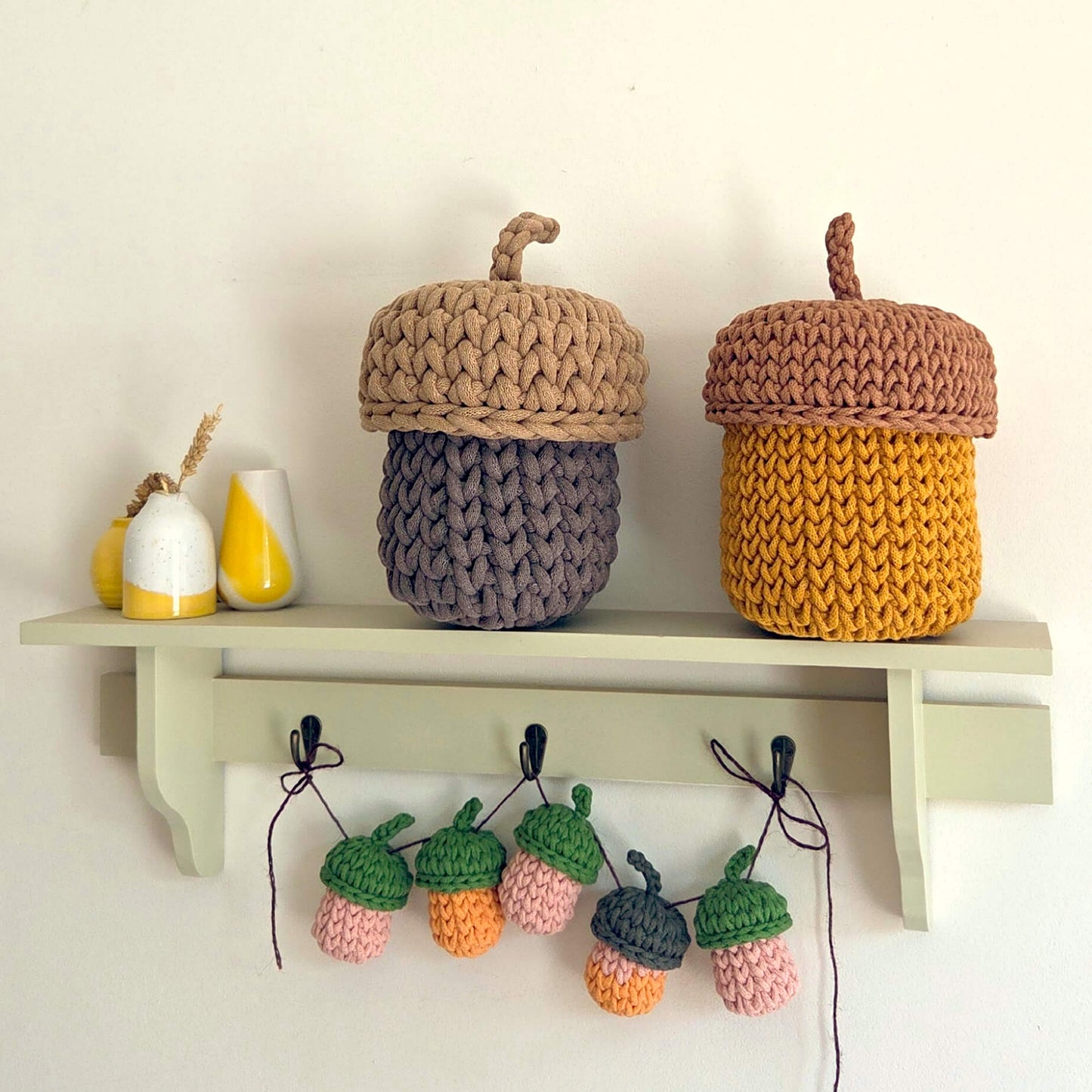 Two crocheted acorn-shaped storage baskets displayed on a light green shelf, with matching hanging acorns below on hooks against a white wall.
