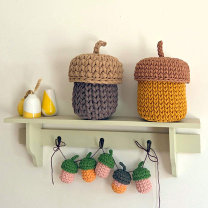 Two crocheted acorn-shaped storage baskets displayed on a light green shelf, with matching hanging acorns below on hooks against a white wall.