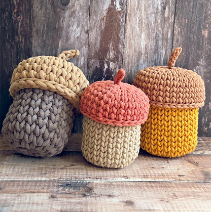 Three crocheted acorn-shaped decorative containers or baskets in autumn colors, arranged on a rustic wooden surface against a weathered wood plank background.