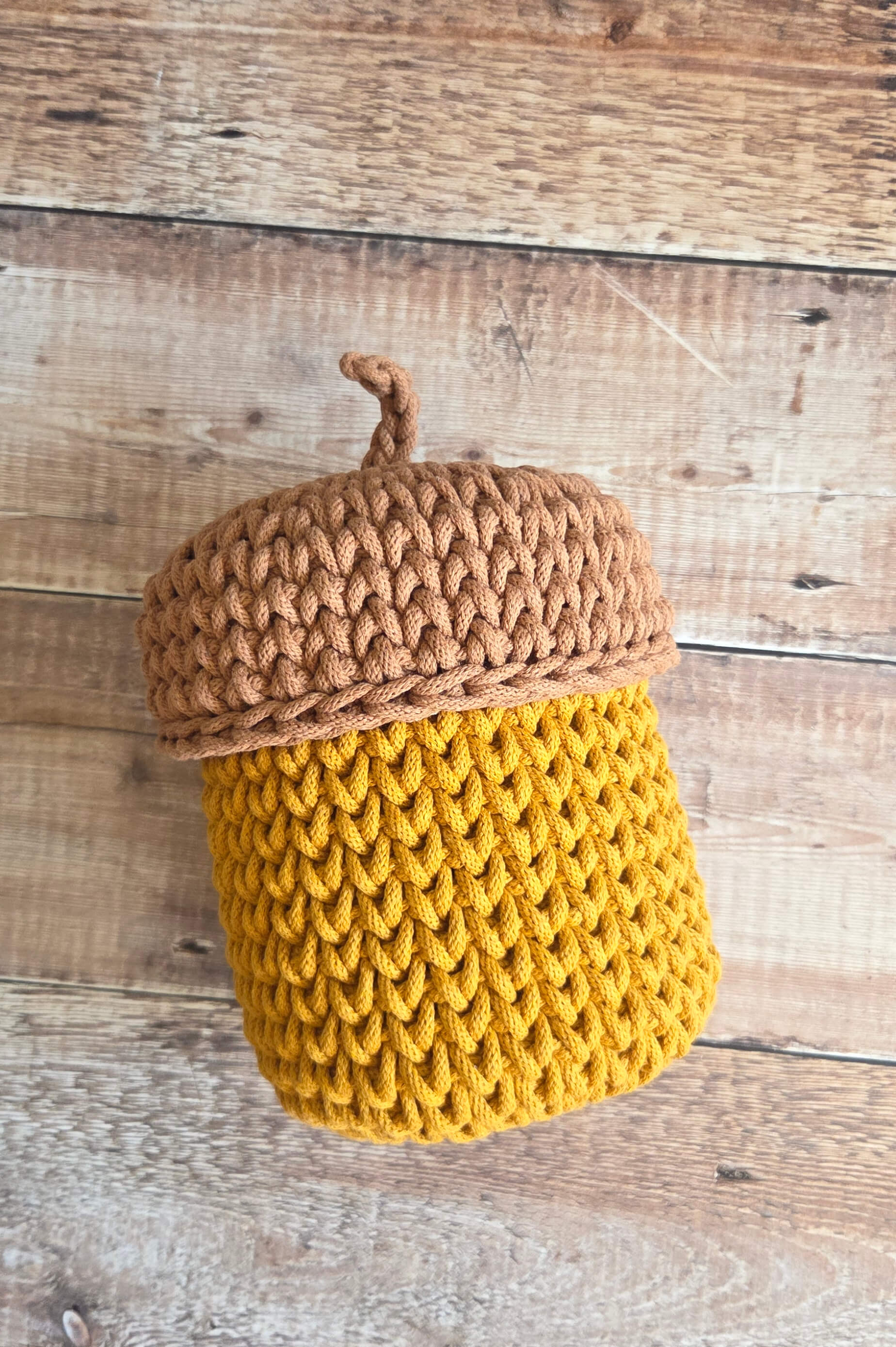 A crocheted acorn-shaped storage basket with a mustard yellow body and a tan-brown textured lid, displayed on a rustic wooden plank surface.