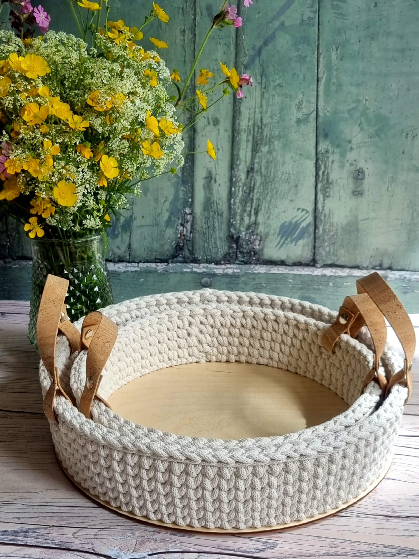 Woven pet bed with brown leather straps next to a vase of flowers on a wooden surface.