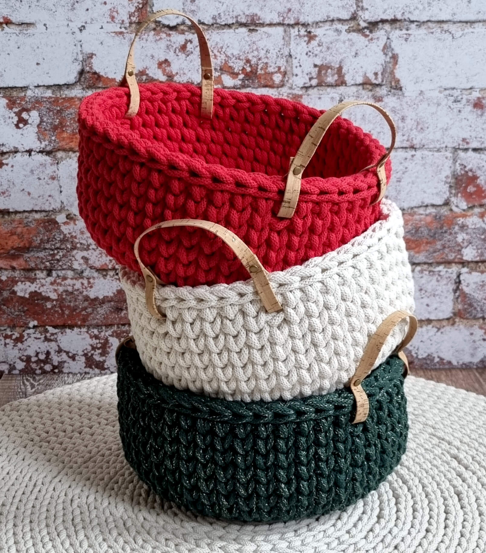 Three crocheted baskets in red, white, and green stacked on a textured surface with a brick wall background.