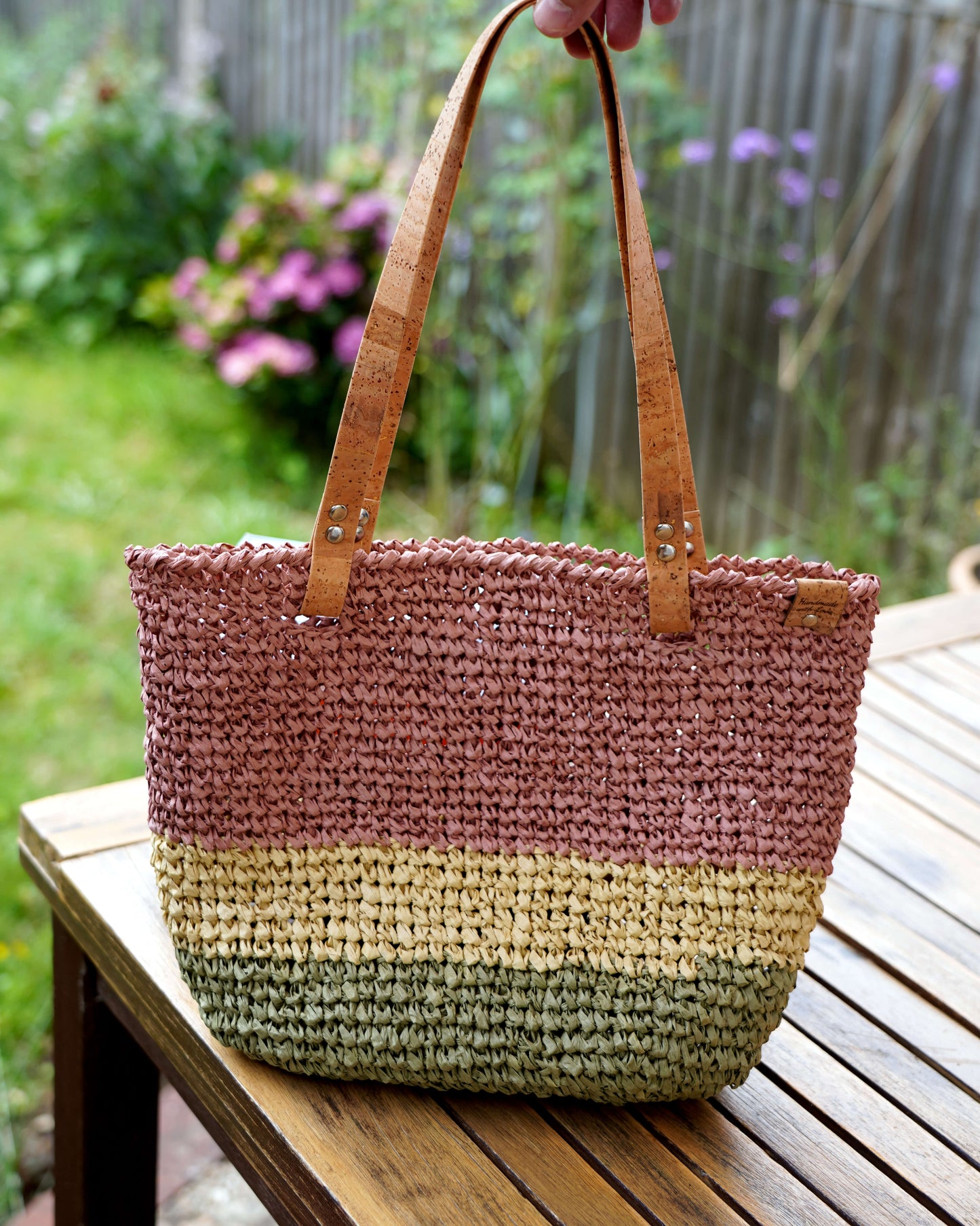 Multicolored woven bag with leather handles on a wooden bench outdoors.