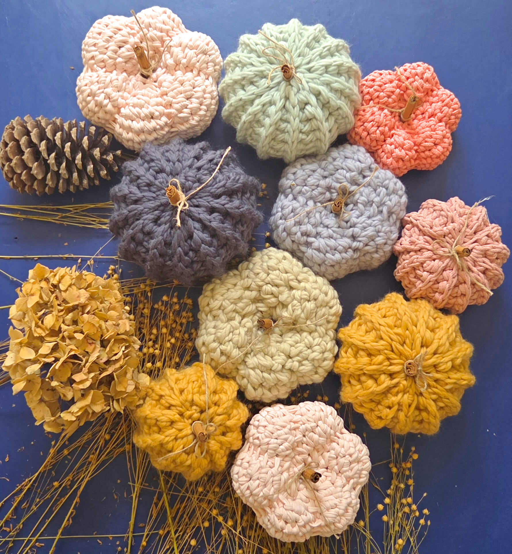 A collection of crocheted pumpkins and dried botanicals arranged on a dark blue background. The pumpkins are made from chunky yarn in pastel peach, sage green, coral, charcoal, light blue-gray, cream, mustard yellow, and light pink.