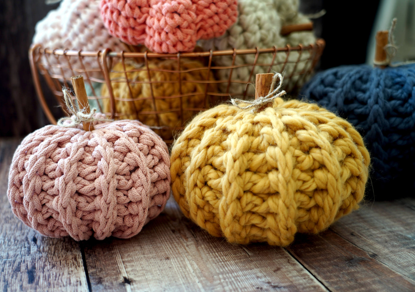 Three chunky knit crocheted pumpkins in dusty pink, mustard yellow, and navy blue arranged on a rustic wooden surface, with a wire basket of more knit pumpkins in the background.