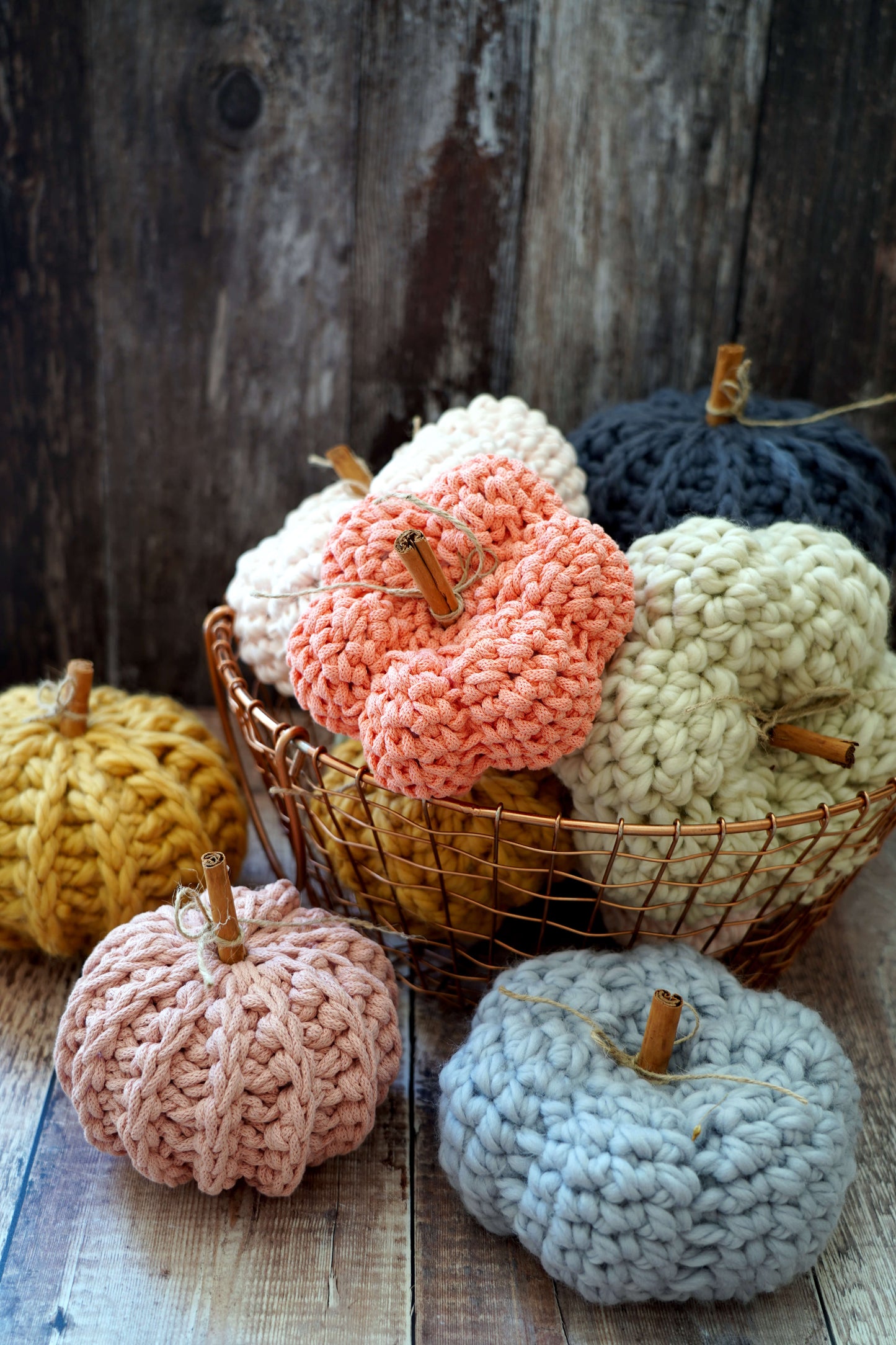 A group of chunky crocheted decorative pumpkins in multiple colors, including yellow, peach, cream, navy, pink, and light blue, arranged on a rustic wooden surface with a wire basket holding the central cluster.