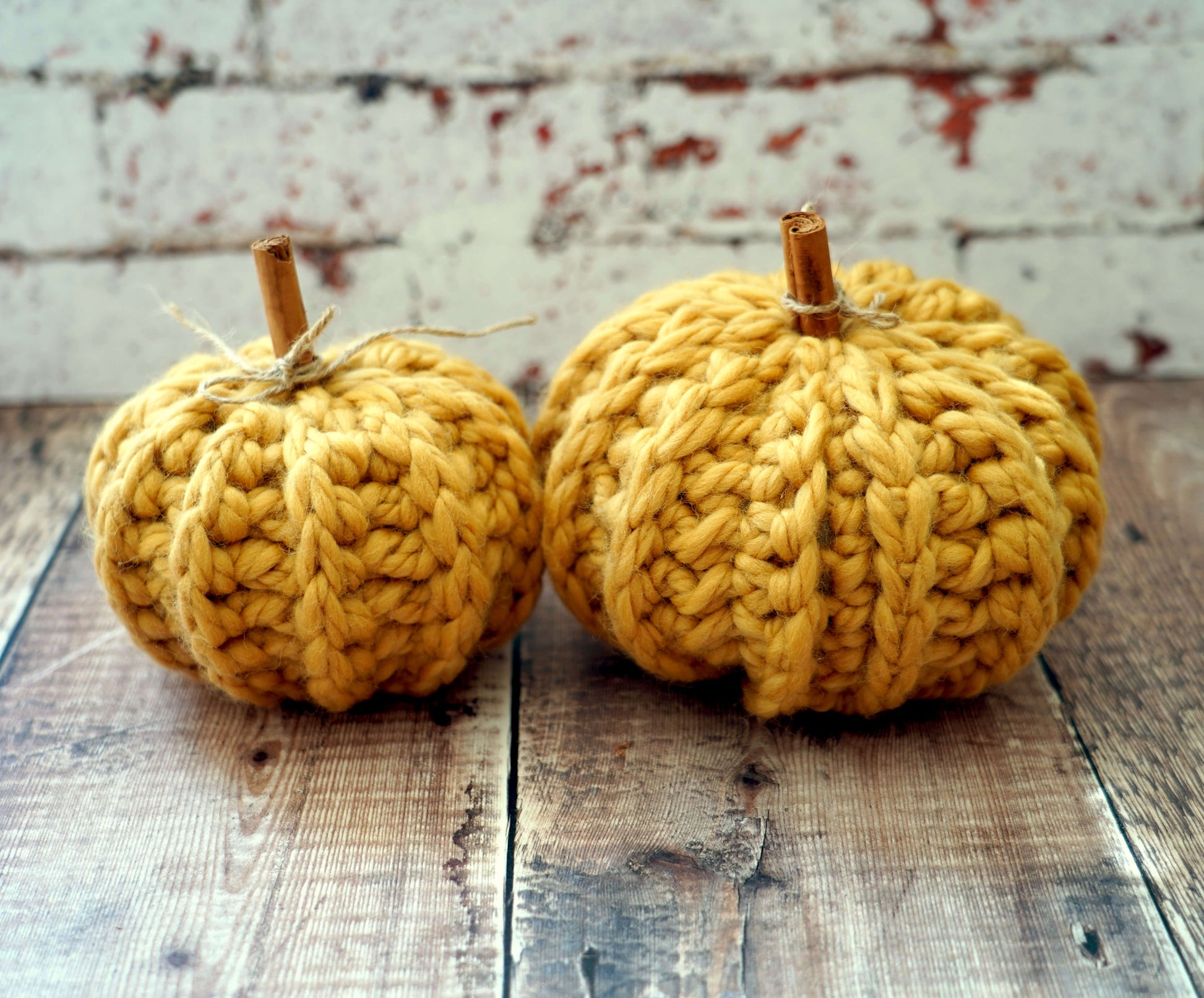 Two small crocheted pumpkins in a warm mustard yellow color, displayed on a rustic wooden surface against a distressed white brick wall background.