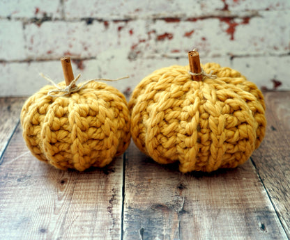 Two small crocheted pumpkins in a warm mustard yellow color, displayed on a rustic wooden surface against a distressed white brick wall background.