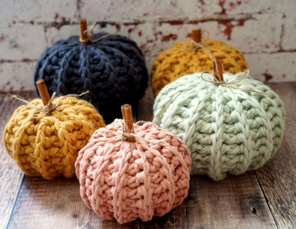 Five chunky crocheted pumpkins in navy, mustard yellow, sage green, and dusty pink, arranged on a rustic wooden surface against a distressed white brick wall.