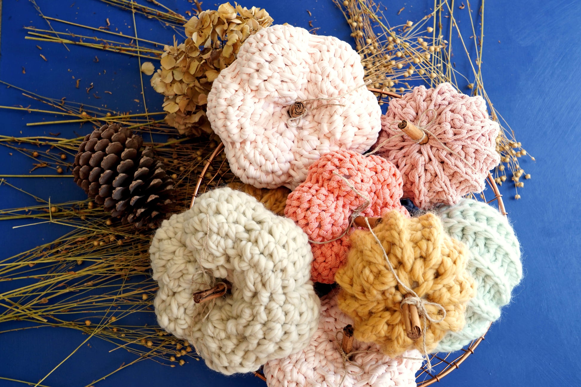 A collection of handmade crocheted pumpkins in various pastel and autumn colors, including cream, pink, peach, mustard, and mint, arranged in a decorative wreath with pinecones and dried grass on a solid blue background.