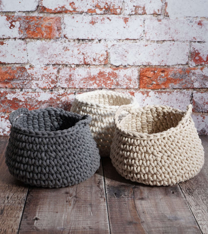 Three knitted baskets in gray, white, and beige on a wooden surface with a brick wall background.