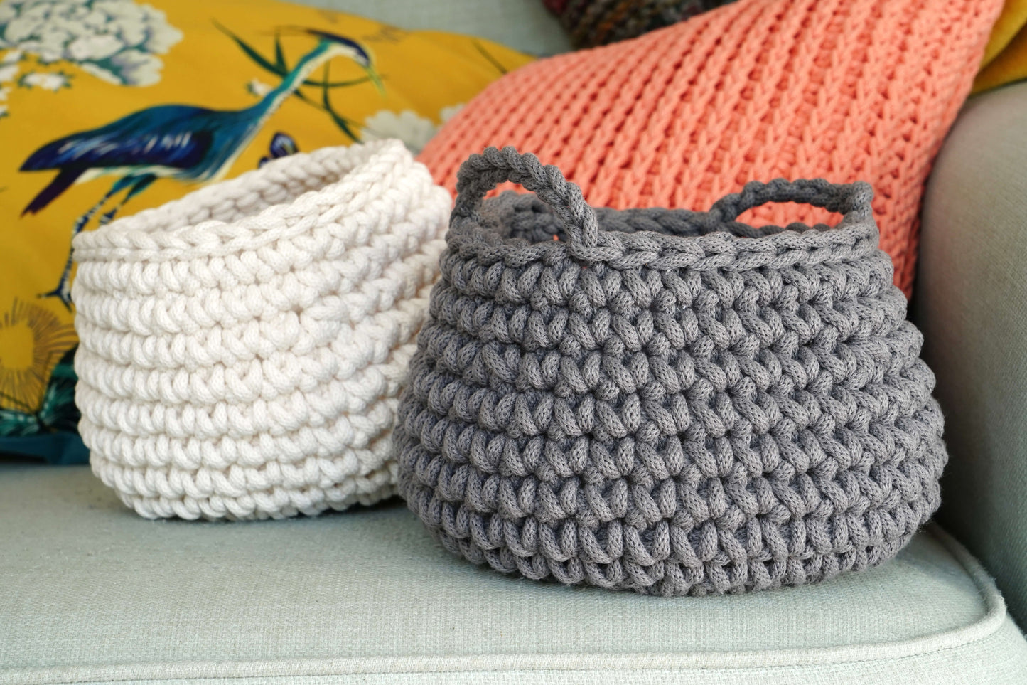 Two knitted baskets, one white and one gray, on a light-colored surface with colorful pillows in the background.