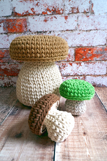 Four crocheted mushroom-shaped items in natural beige, brown, green, and cream colors, arranged on a rustic wooden surface against a distressed white and red brick wall.