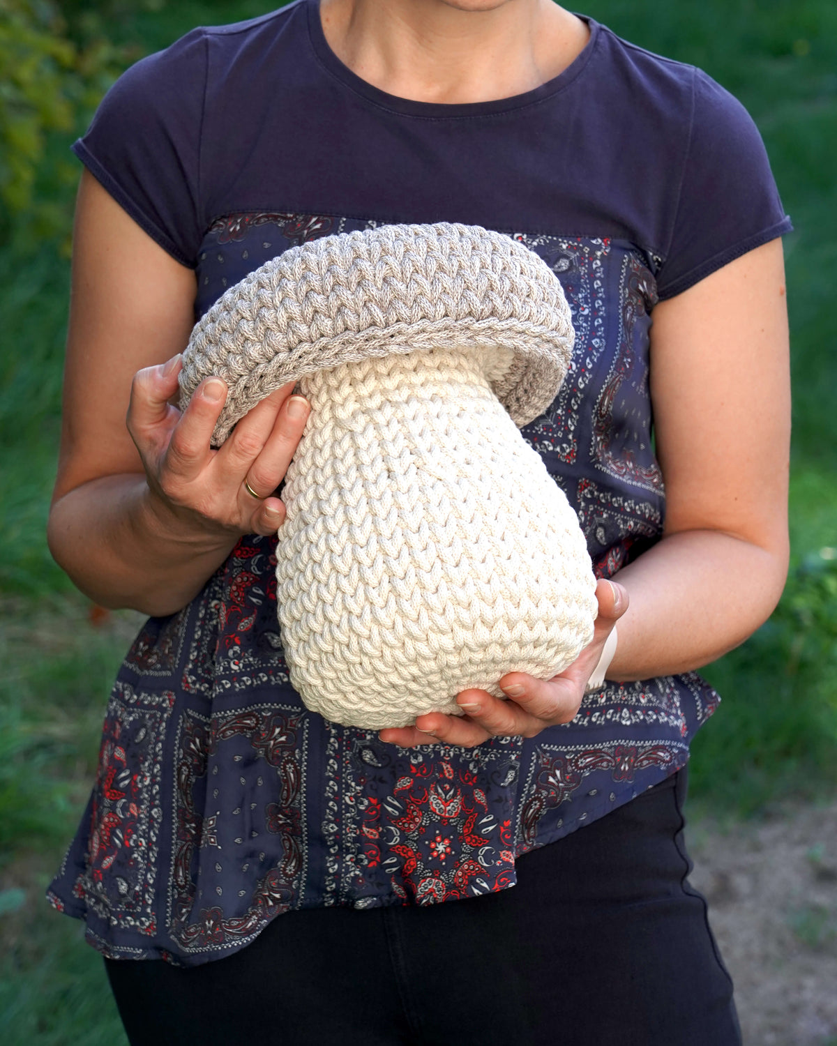 A person outdoors holding a large, chunky hand-knit mushroom decoration. The person is wearing a navy blue t-shirt and a patterned blouse, with green foliage blurred in the background.