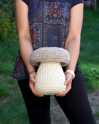 A person outdoors holding a chunky knit mushroom-shaped decorative item in cream-colored yarn. The person is wearing a navy blue patterned top and black pants, with a green garden background.