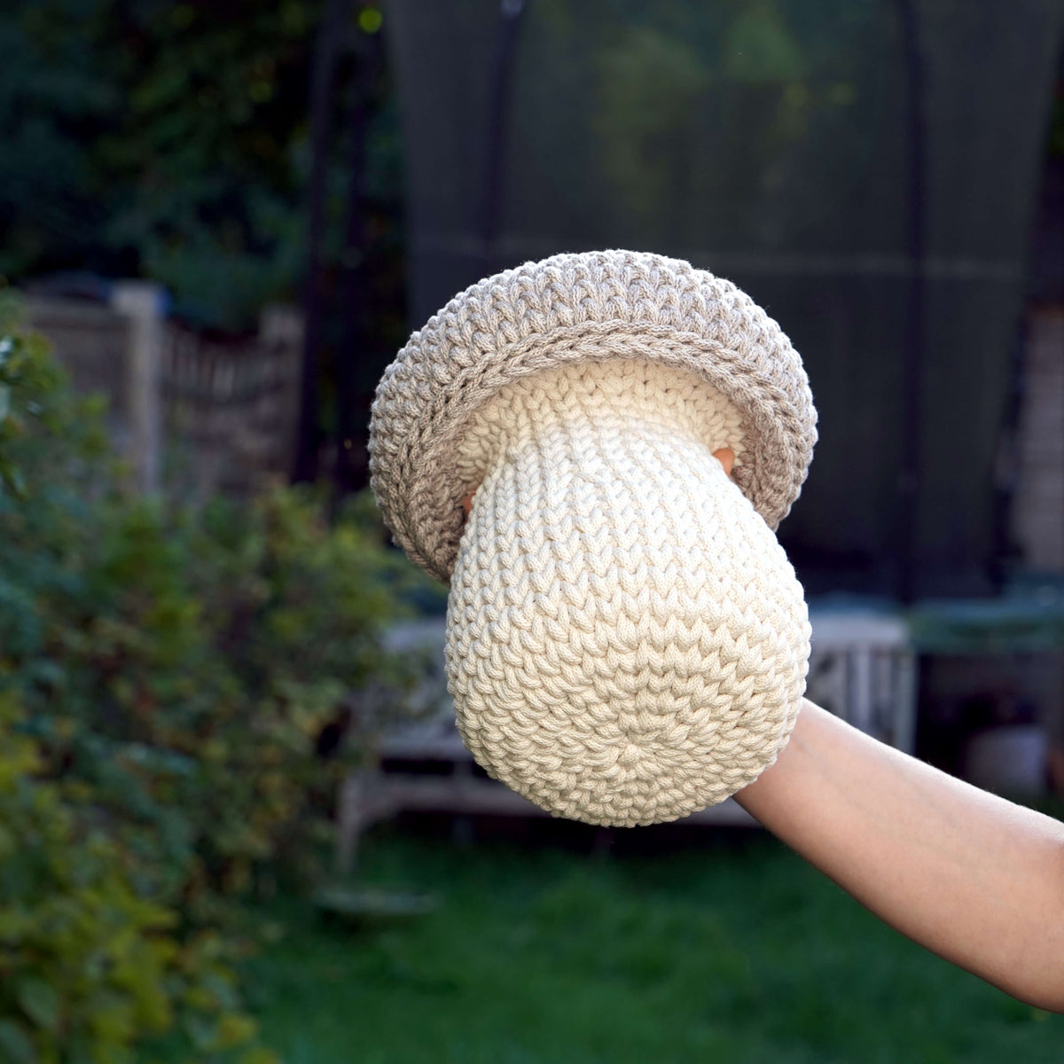 A hand wearing a chunky knit mushroom-shaped glove, photographed outdoors with a blurred garden background.