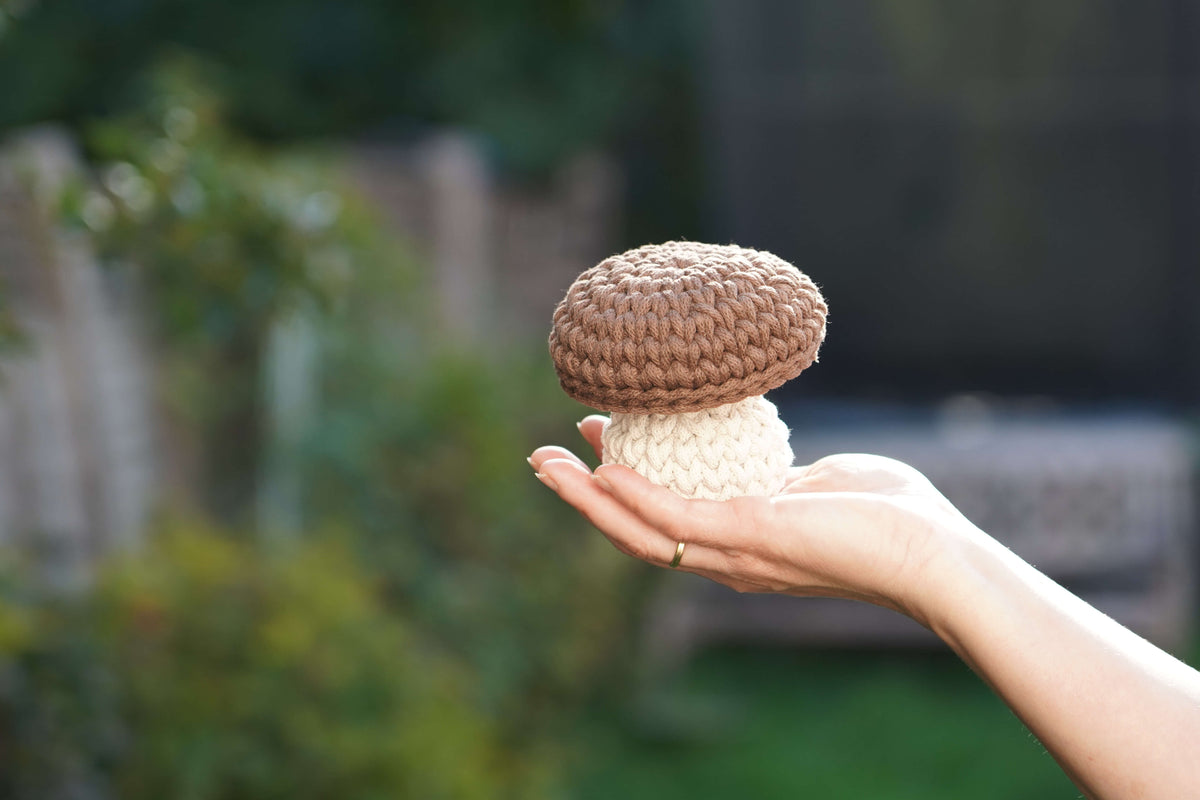 A hand holding a small crocheted mushroom toy outdoors, with a blurred garden background.