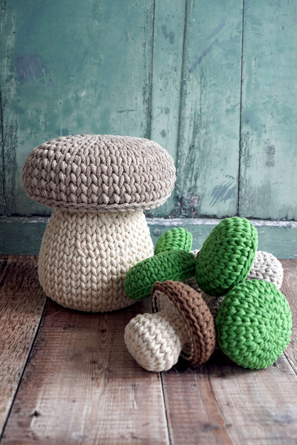 Handmade crocheted mushroom-shaped items displayed on a rustic wooden surface against a distressed teal-green wooden wall. A large beige and cream mushroom sits on the left, while several smaller green, brown, and cream crochet mushrooms are arranged in front.