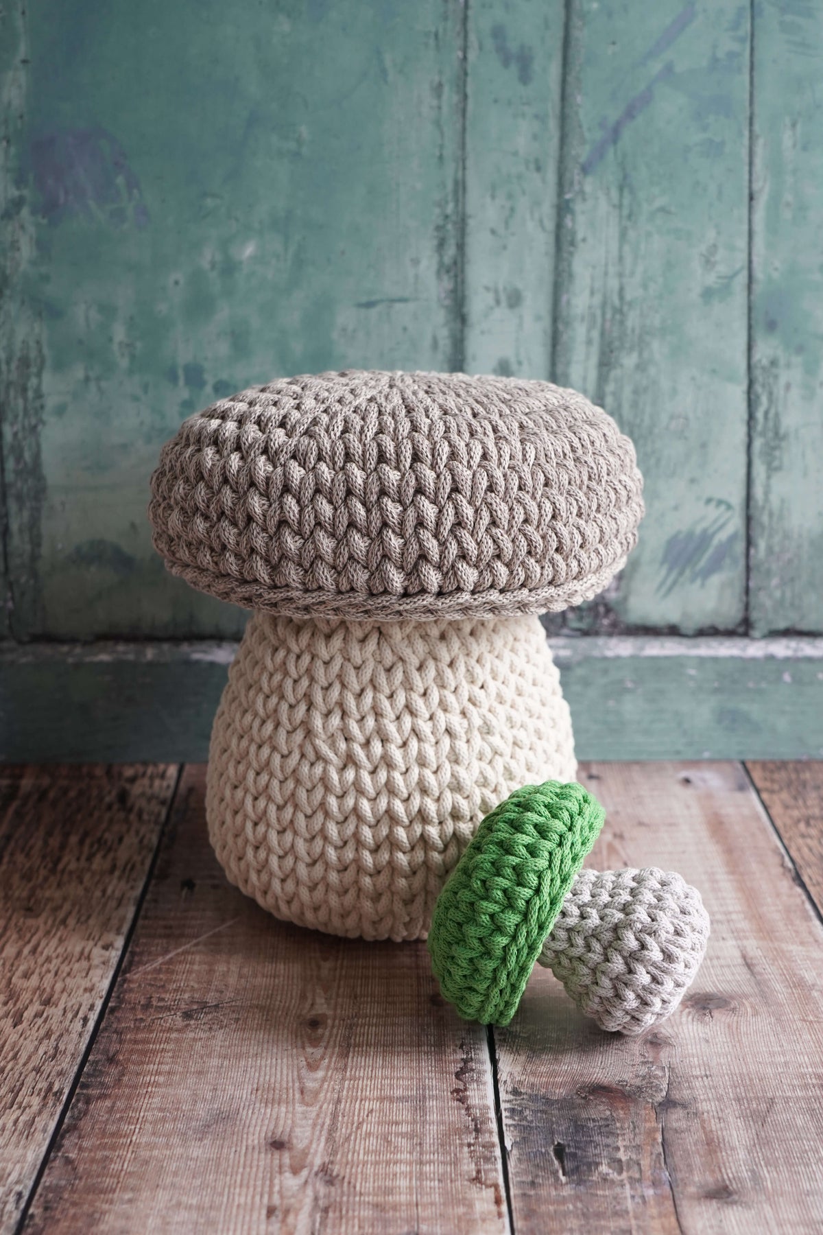 A handmade mushroom-shaped pouf stool made of chunky knit yarn, shown alongside two smaller crochet mushroom toys on a rustic wooden floor against a distressed teal-green wooden wall.