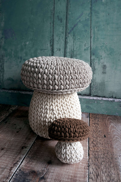 Three hand-crocheted mushroom-shaped poufs or stools in neutral tones, arranged on a rustic wooden floor against a distressed teal-green wooden wall.