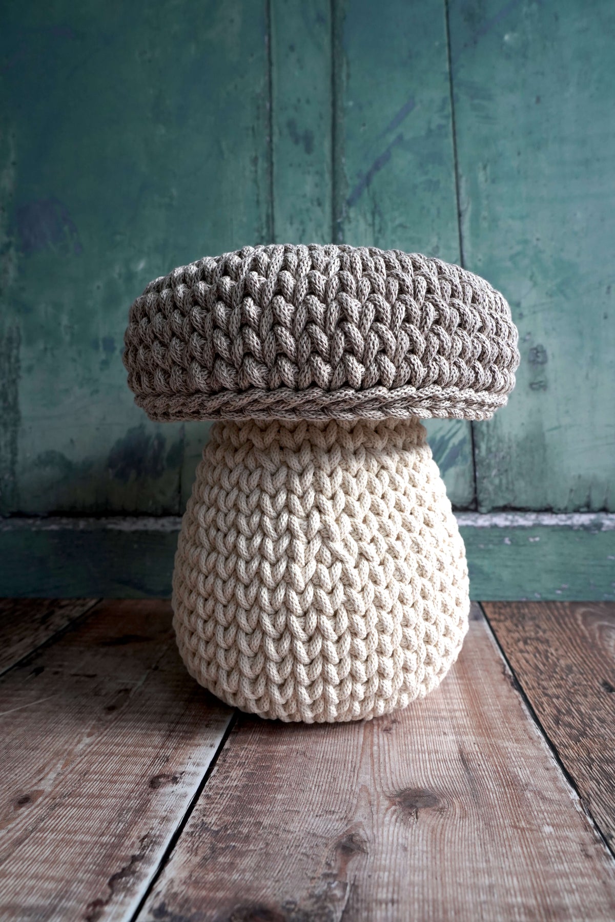 A mushroom-shaped knitted pouf stool with a textured beige cap and a cream-colored stem, set on a rustic wooden floor against a distressed teal-green wooden wall.