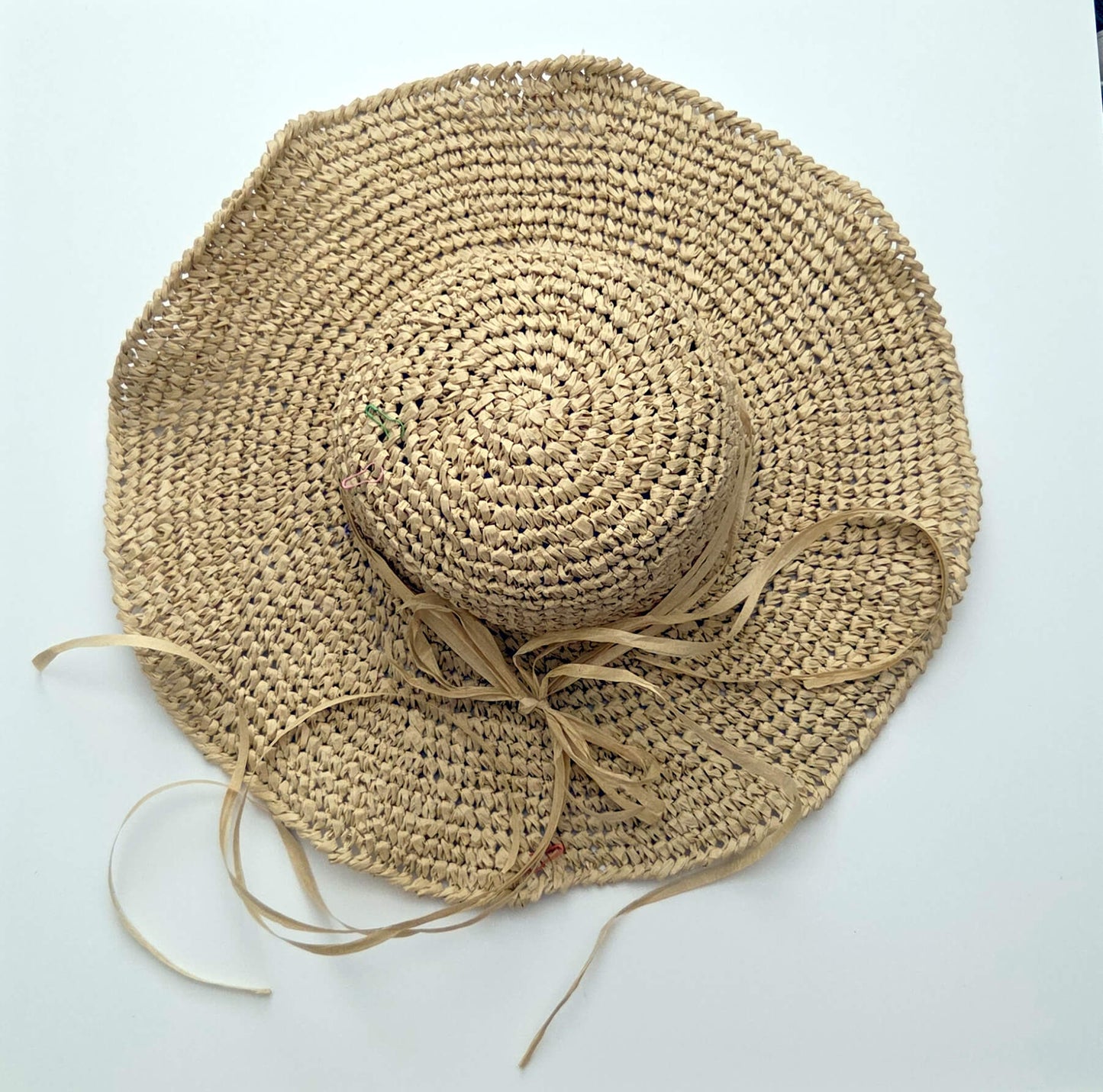 A natural straw sun hat with a wide brim, a small crown, and long ribbon ties, laid flat on a white background.