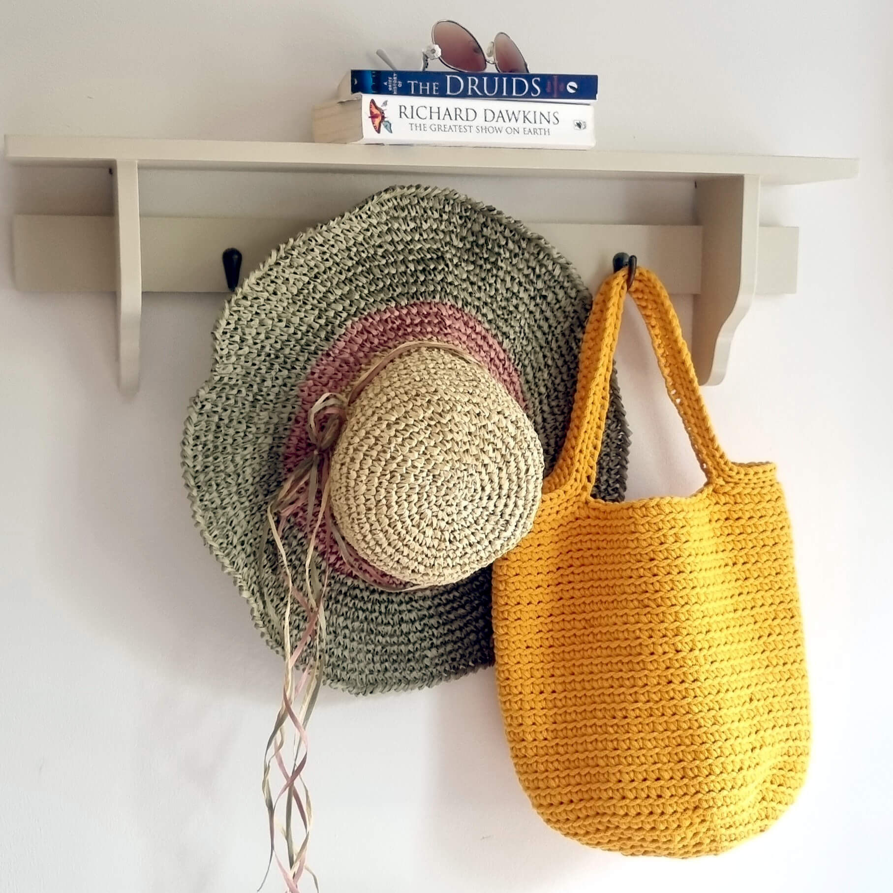A yellow crocheted tote bag hangs on a white wall hook next to a large woven hat in sage green and cream with a dusty pink band, with a white shelf above holding books and sunglasses.