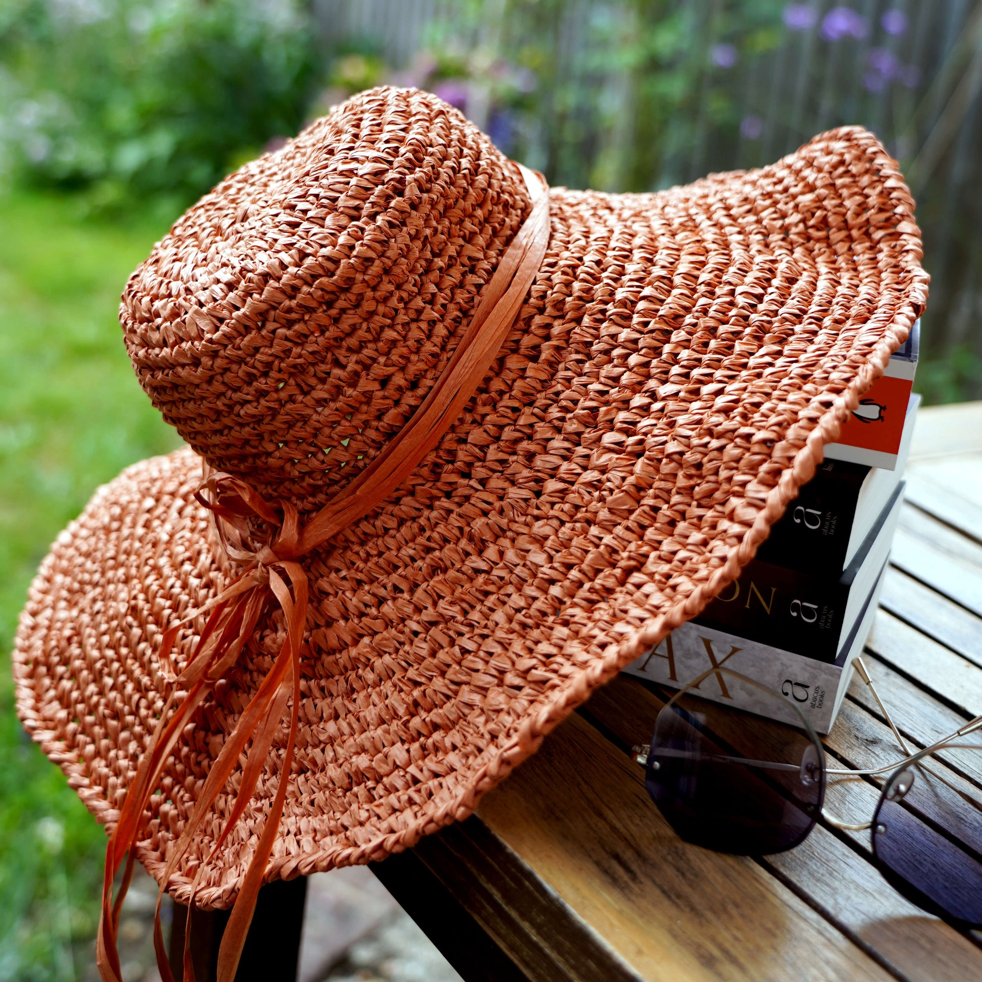 A woven wide-brim sun hat in a warm terracotta or rust orange-brown color, tied with a matching ribbon, resting on a wooden table outdoors with sunglasses and books nearby.