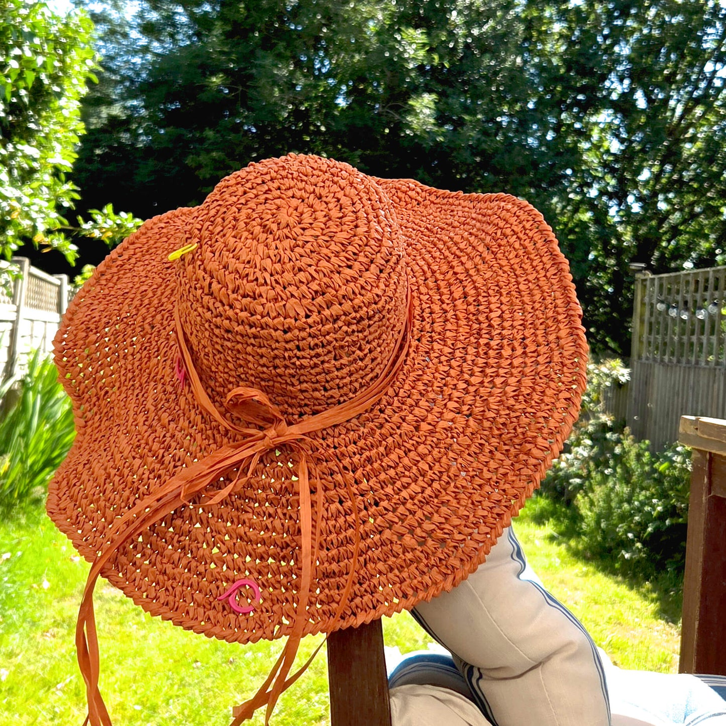 A wide-brimmed woven straw sun hat in a warm terracotta orange color, featuring a matching orange ribbon tie and small pink decorative accents, photographed outdoors on a wooden stand in a backyard setting.