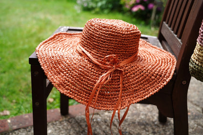 A wide-brimmed woven sun hat in a warm terracotta orange color, tied with a matching ribbon, resting on a dark wooden bench in an outdoor garden setting.