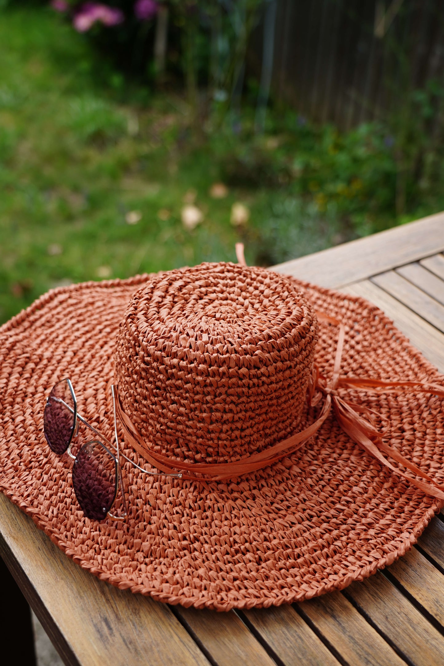 A terracotta-colored woven sun hat with a matching ribbon band and attached sunglasses, resting on a wooden table with a blurred garden background.