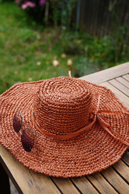 A terracotta-colored woven sun hat with a matching ribbon band and attached sunglasses, resting on a wooden table with a blurred garden background.
