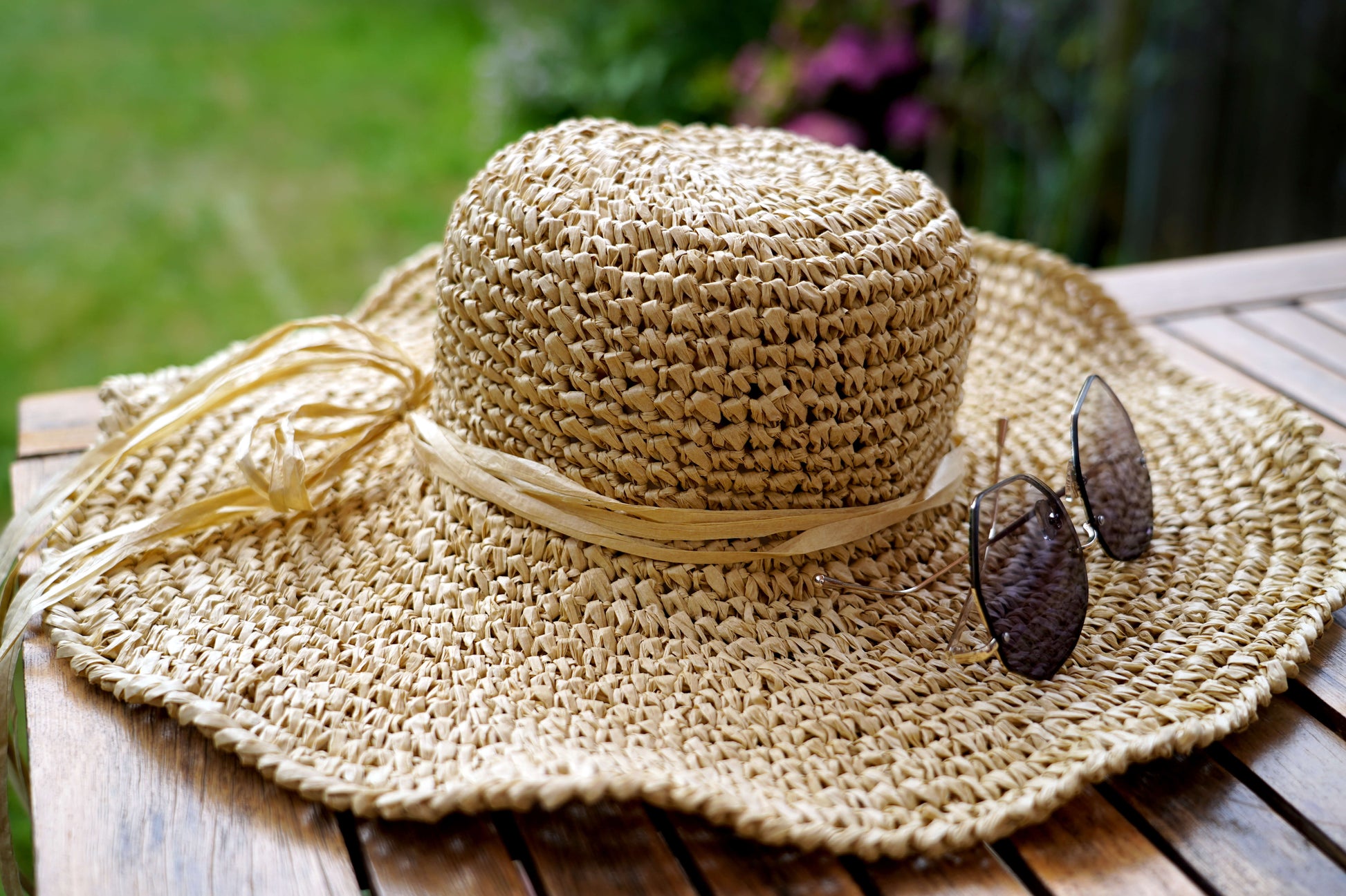 A natural straw sun hat with a wide brim, a matching ribbon band, and a decorative raffia tie, resting on a wooden table outdoors with sunglasses placed on the brim.