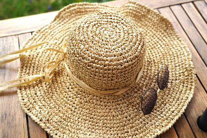 A natural straw sun hat with a wide brim and a matching ribbon band, styled on a wooden table with a pair of sunglasses resting on the brim.