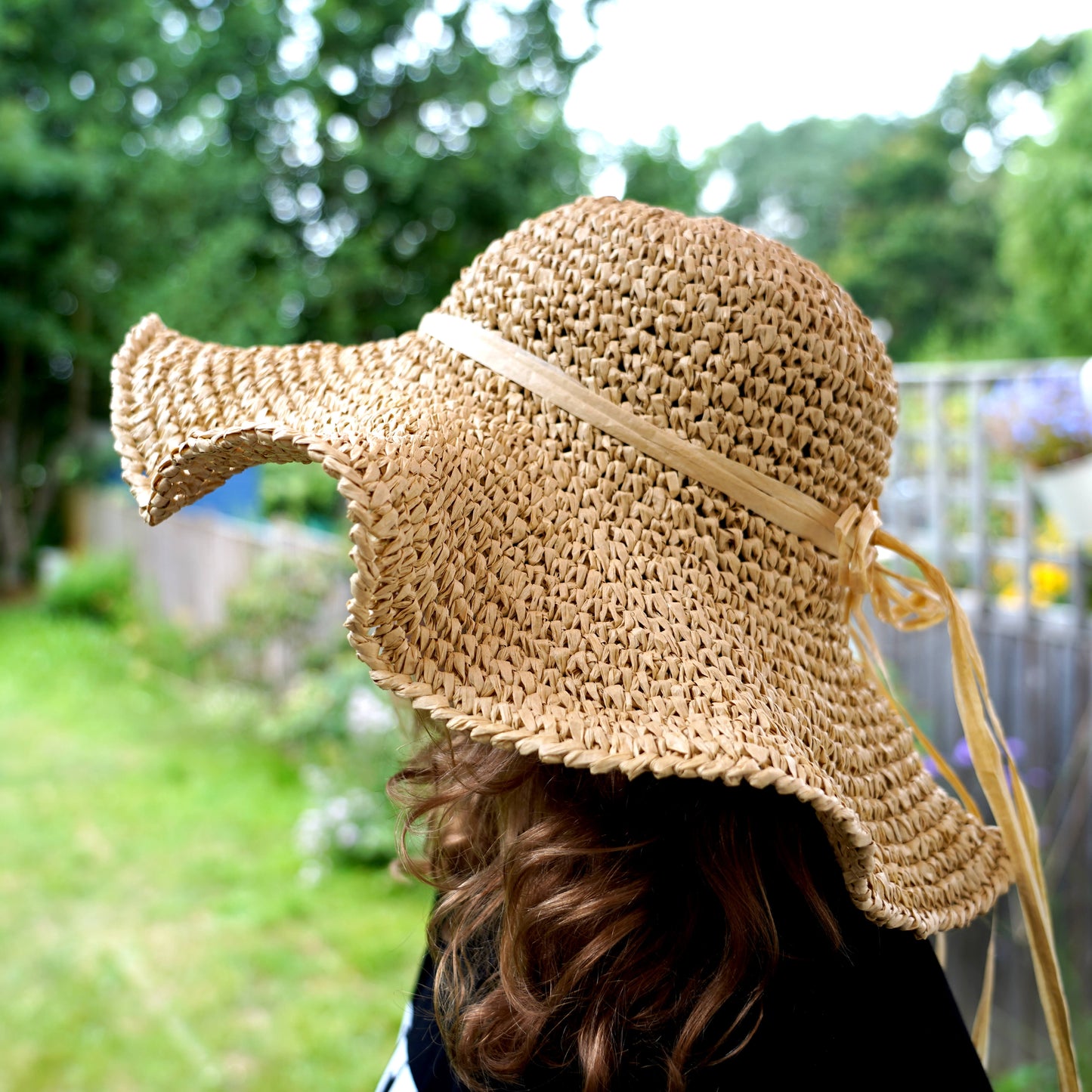 A straw sun hat with a wide brim and ribbon tie, worn by a person in an outdoor garden setting.