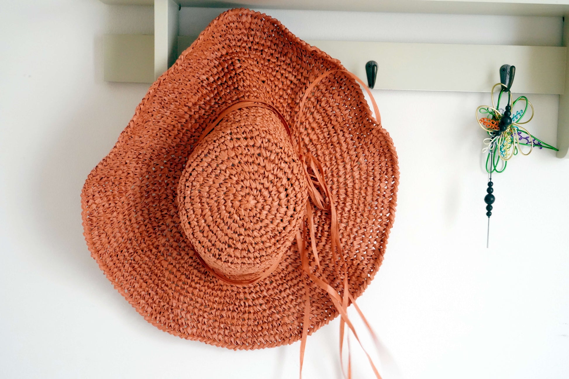 A handwoven terracotta orange straw hat with a matching ribbon tie, hanging from a white wall hook.