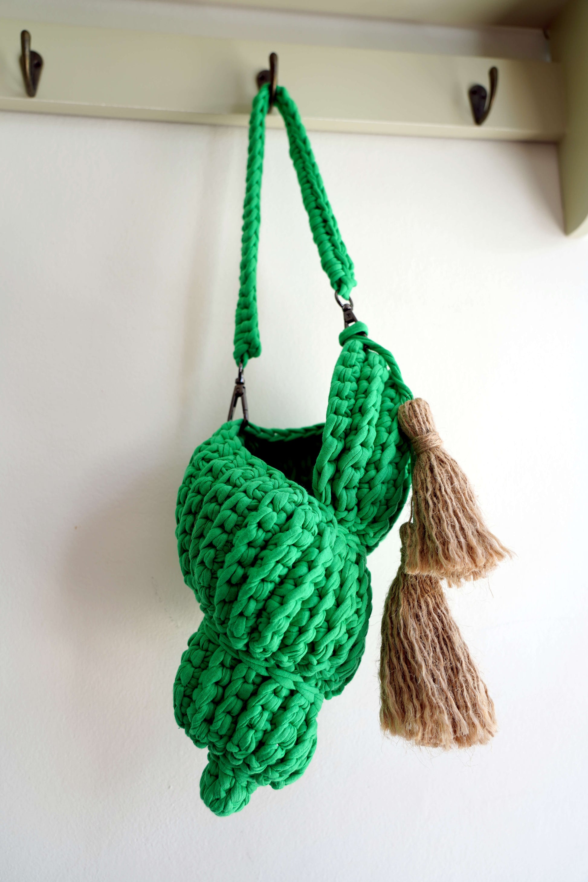 Green knitted bag with tassels hanging on a hook against a white wall.
