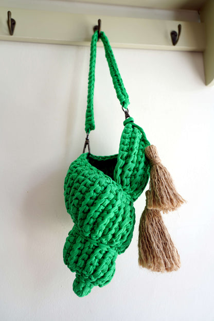 Green knitted bag with tassels hanging on a hook against a white wall.